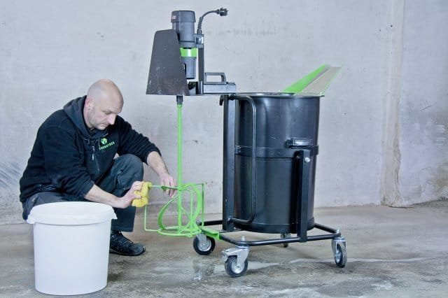 Cleaning mixing station A man in a black hoodie is using a green mixing tool next to a large black container on wheels, with a small white bucket on the floor.
